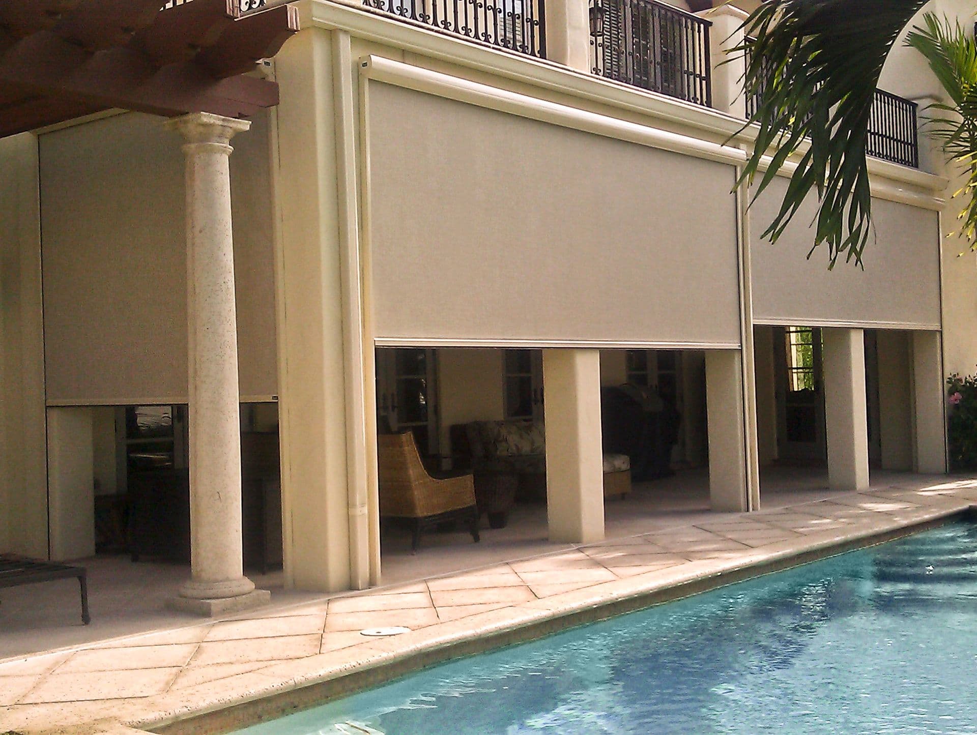 Poolside patio with retractable awnings covering glass doors and windows of a residential building, with stone column and palm plant visible