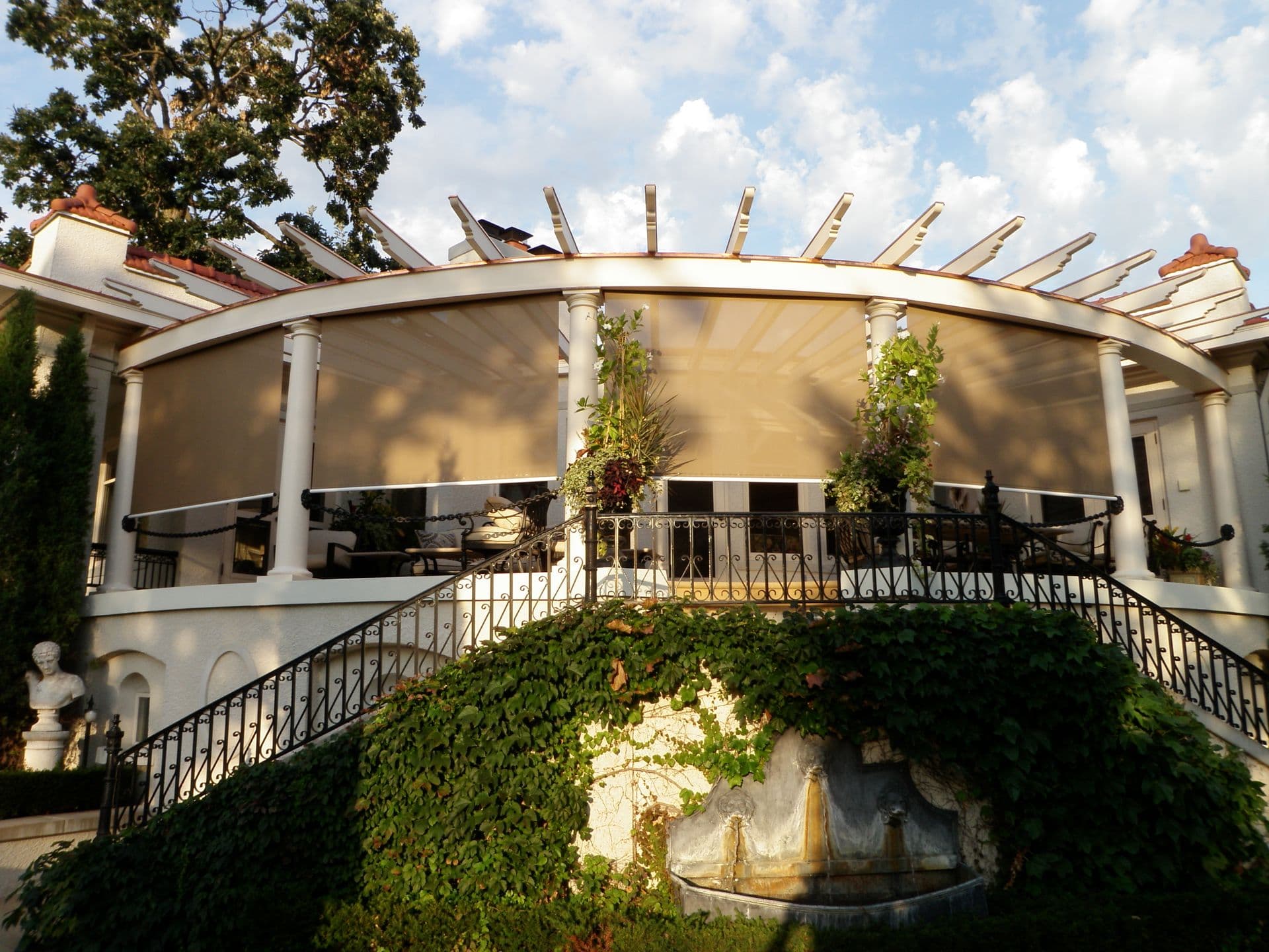 Curved pergola with retractable shade screens on residential terrace, featuring white columns, ornamental railing, and climbing ivy below