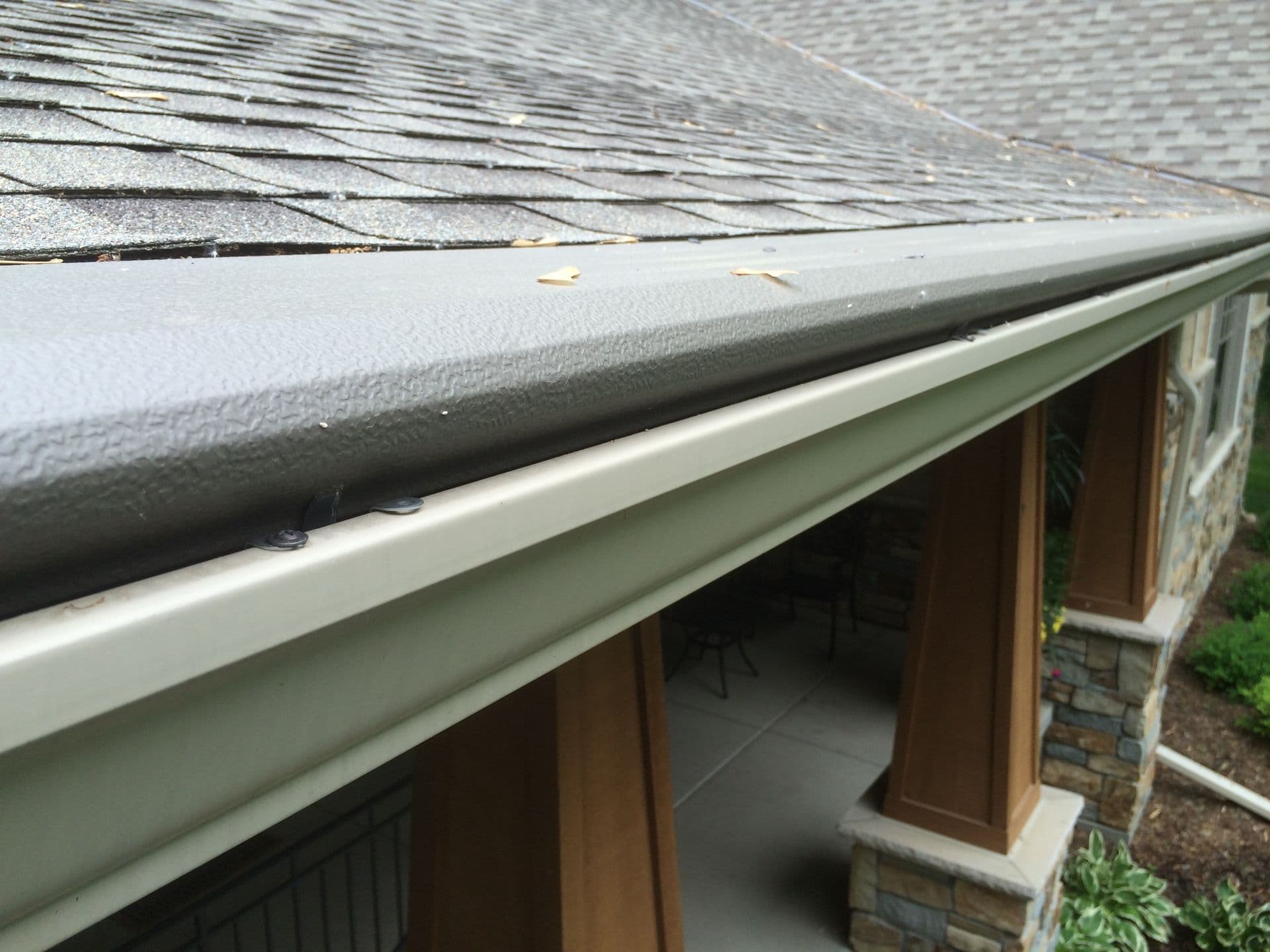 Gray gutter guard covering white aluminum gutter below asphalt shingle roof with debris and fallen leaves visible