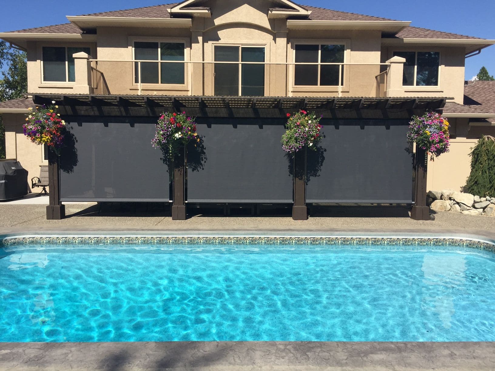 Two-story beige house with retractable shade screens over patio area, decorative hanging flower baskets, viewed from swimming pool in foreground