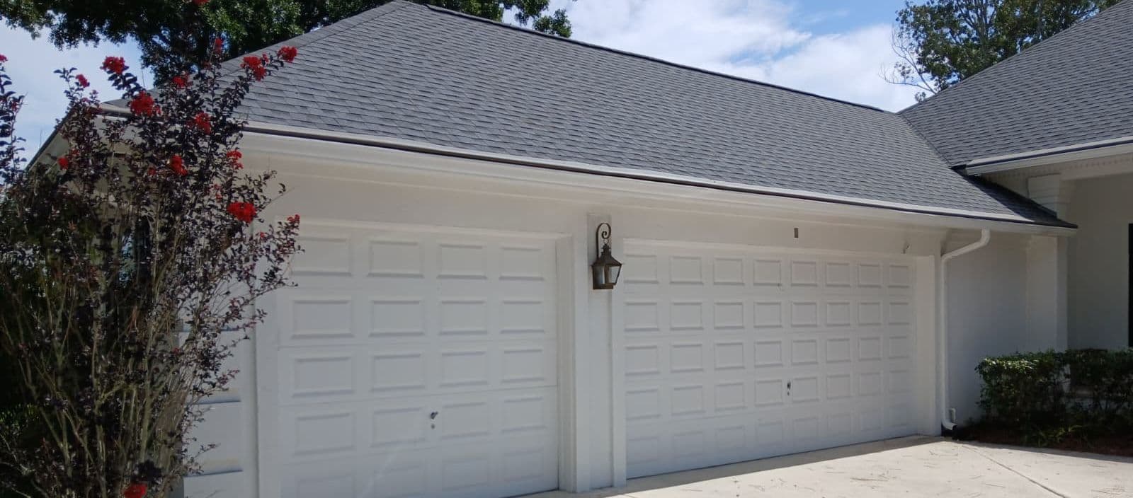 Two-car white garage with dark gray roof, concrete driveway, red flowering vines on left side, and sconce light fixture