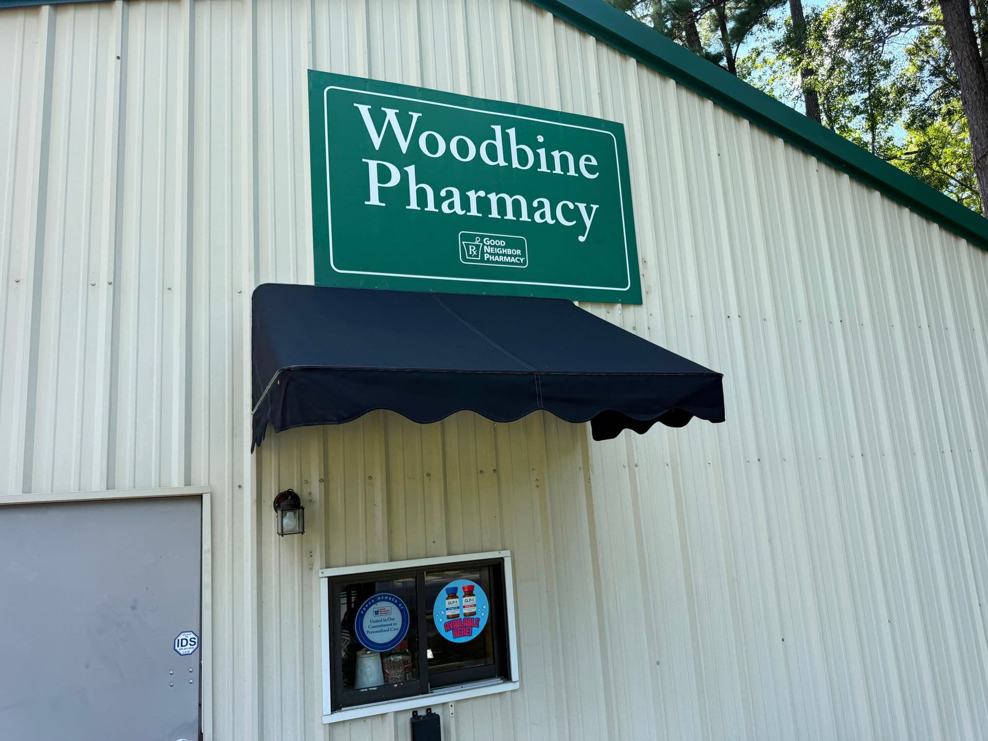 Woodbine Pharmacy storefront with green sign above a black awning and service window on corrugated metal building