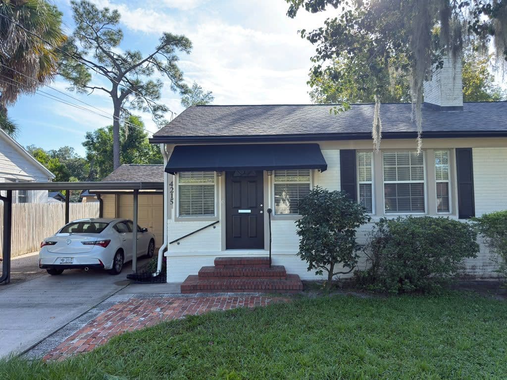 White single-story residential home with black awning over entrance, brick steps, carport with white vehicle, mature trees with Spanish moss, and manicured lawn