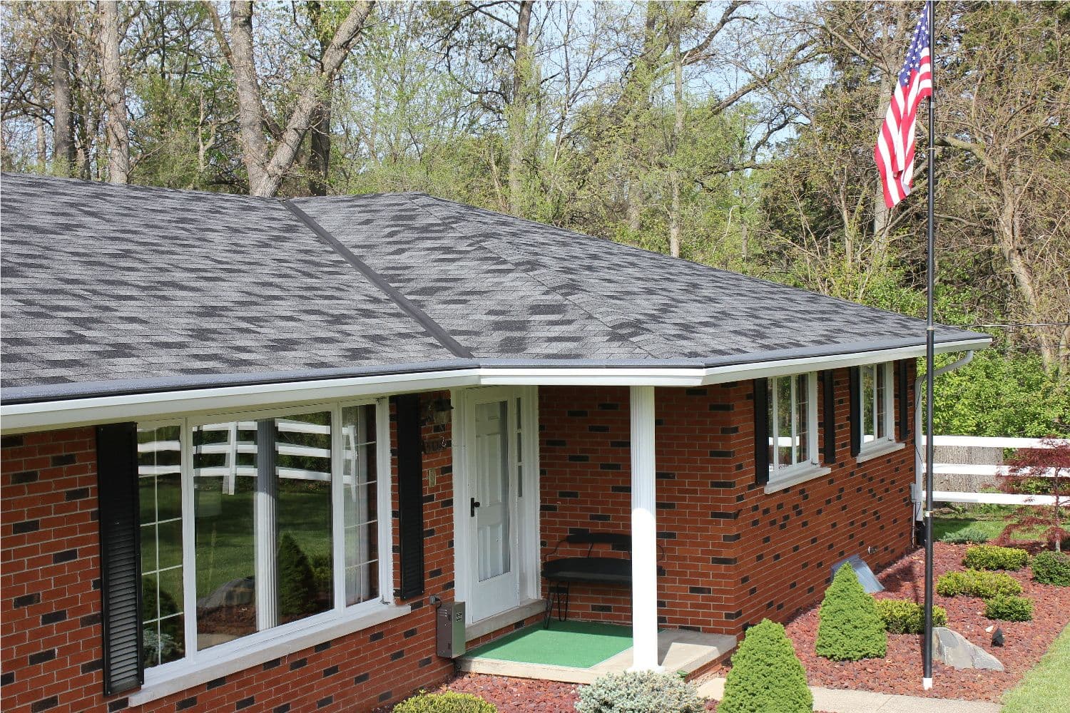 Single-story brick home with black asphalt roof, white trim windows and door, front porch with white columns, American flag on pole, surrounded by mature trees and landscaping