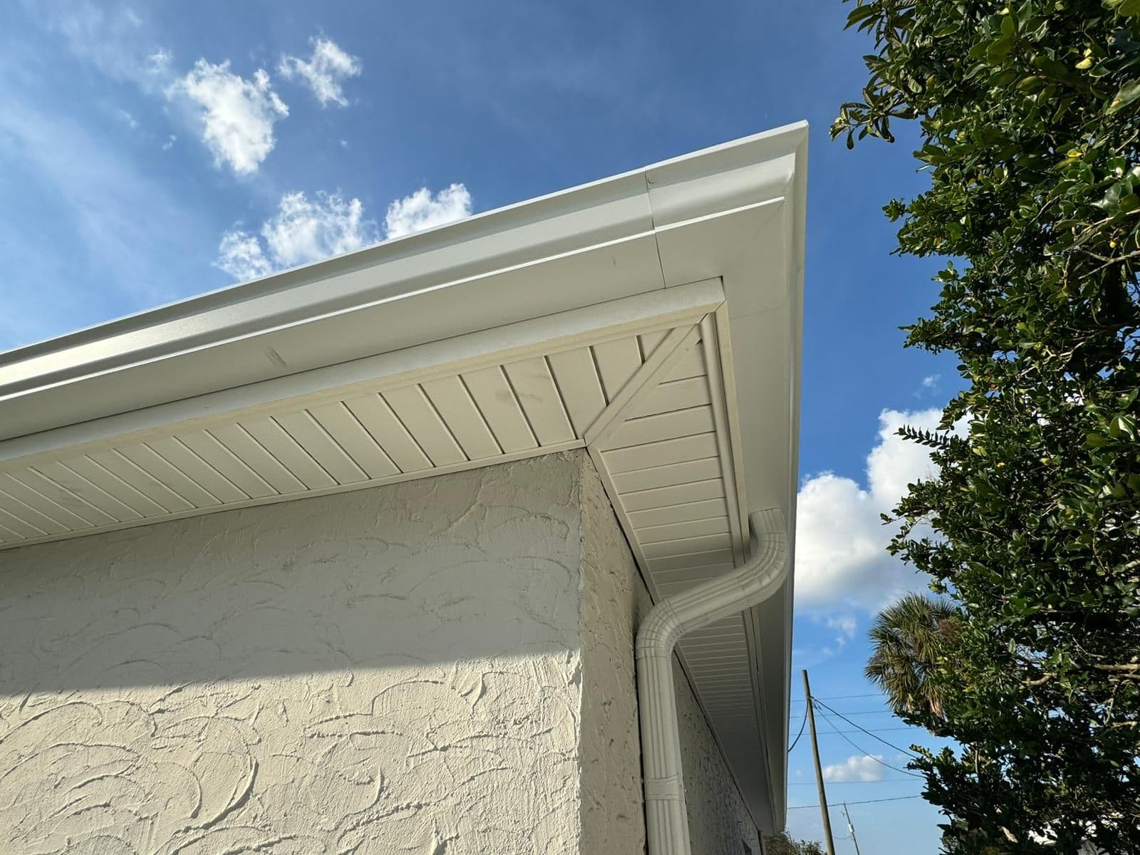 White stucco building exterior with cream soffit and gutter against blue sky, bordered by green foliage
