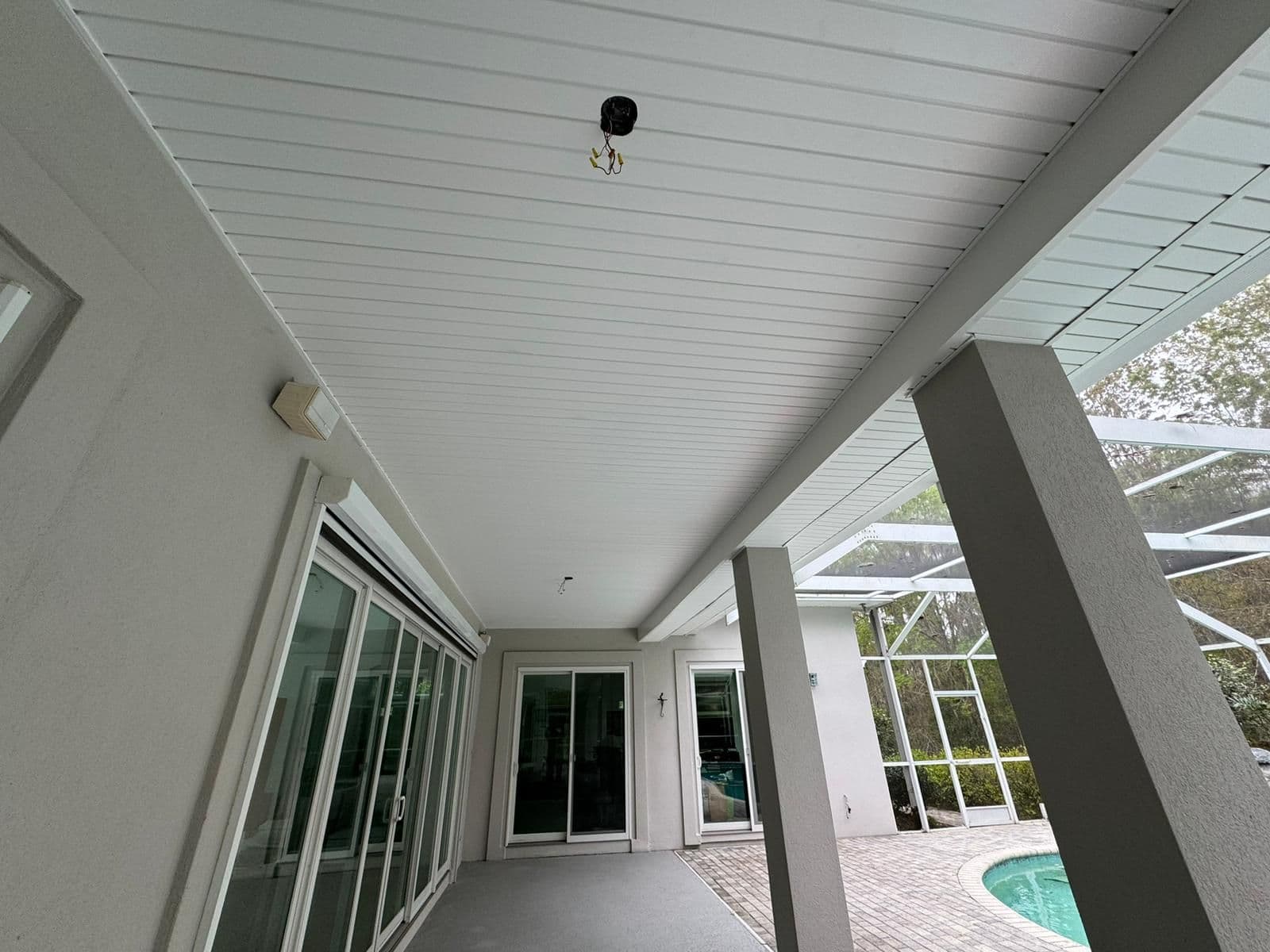 Covered patio with white plank ceiling, concrete support columns, glass sliding doors, and pool area visible through latticed railing on the right