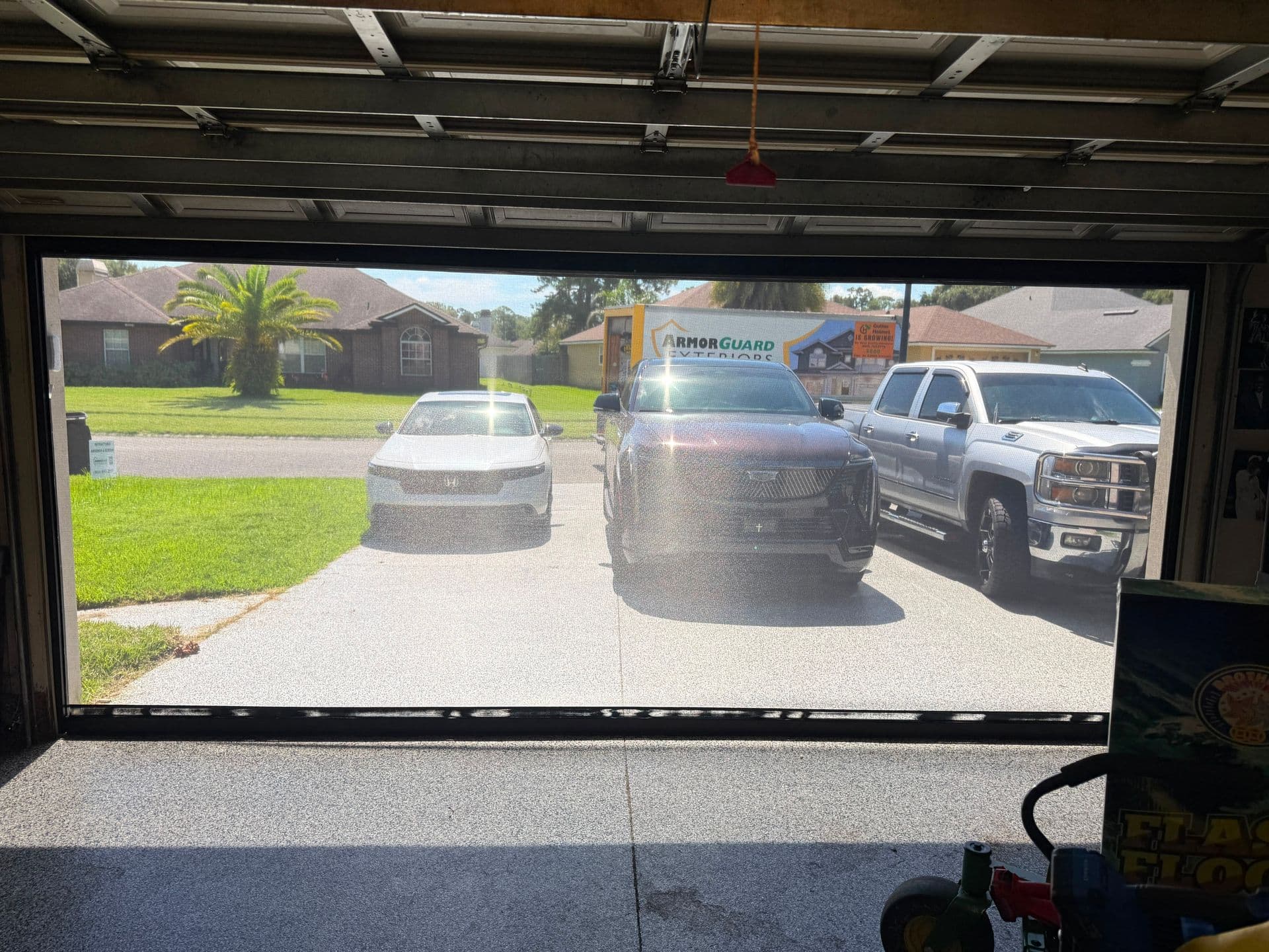 Garage door camera view showing driveway with three vehicles parked in front of suburban house with ArmorGuard sign visible in background