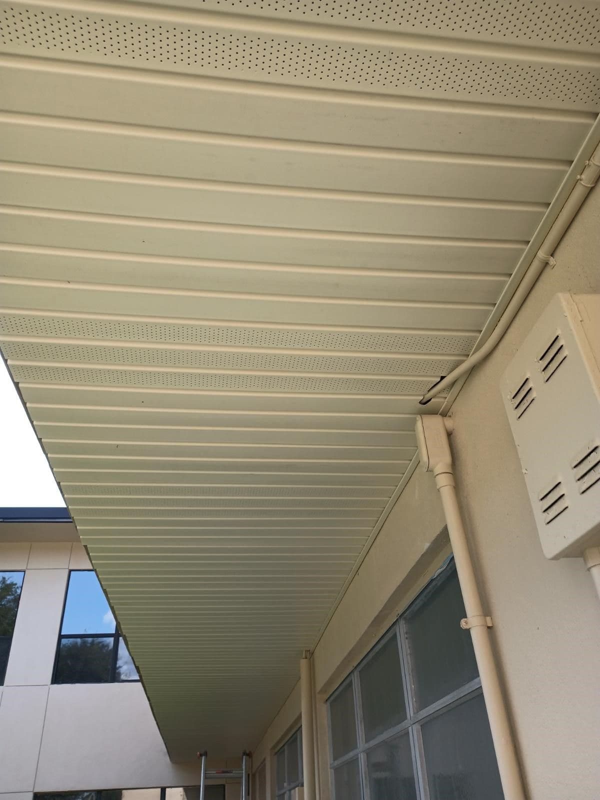 Covered outdoor walkway with metal corrugated ceiling, perforated panels above, downspout, and glass doors along the right side