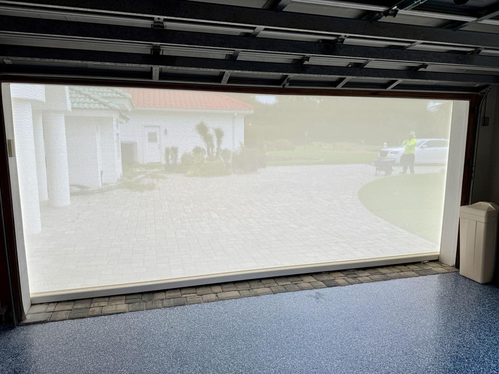 View through garage door window showing driveway, home exterior, landscaping, and parked vehicles