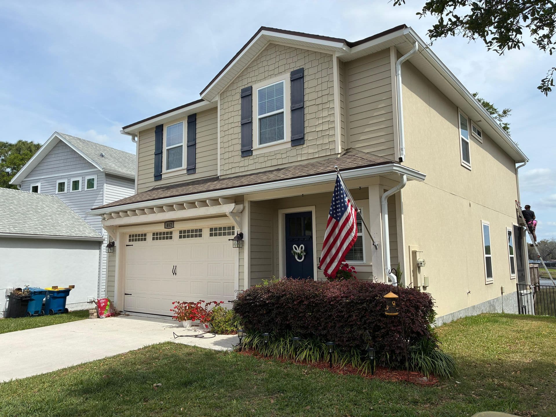 Two-story residential home with tan and cream siding, two-car garage, front porch with American flag, landscaping with dark shrubs and red flowers, neighboring white house visible on left