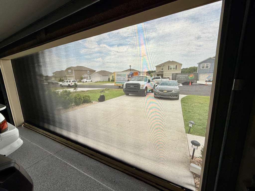 View through garage window showing driveway with parked vehicles, residential neighborhood houses, and green lawn in background